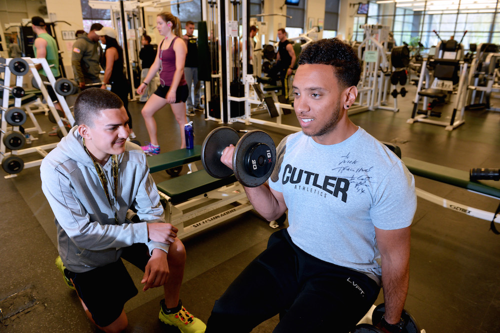 A male student lifting weights while a trainer looks on